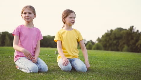 Two-Pretty-Little-Sisters-Talking-Together,-Looking-Around-And-Pointing-Something-While-Kneeling-On-Grass-At-Park-1
