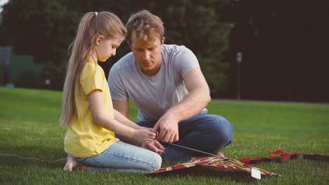 Father-And-Daughter-Fixing-Kite-Sitting-On-A-Green-Grass-In-The-Park