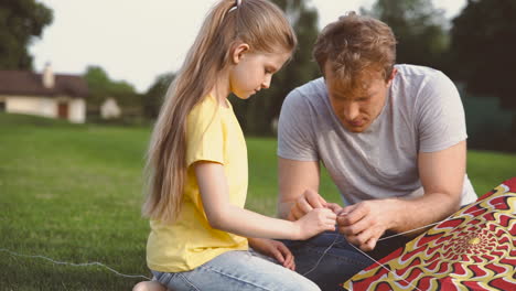 Father-And-Daughter-Fixing-Kite-Sitting-On-A-Green-Grass-In-The-Park-1