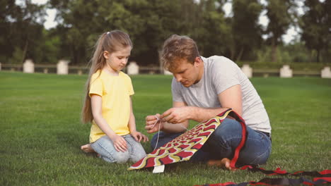 Father-Fixing-Kite-Sitting-On-Meadow-With-His-Little-Daughter