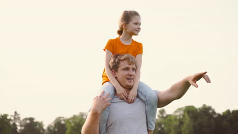 Happy-Father-Carrying-His-Daughter-On-Shoulders-In-A-Park-While-Walking-And-Explaining-Her-Something