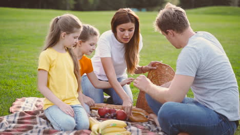 Glückliche-Eltern-Mit-Zwei-Kleinen-Töchtern,-Die-Essen-Aus-Dem-Korb-Nehmen,-Während-Sie-Zusammen-Auf-Der-Grünen-Wiese-Im-Park-Picknicken