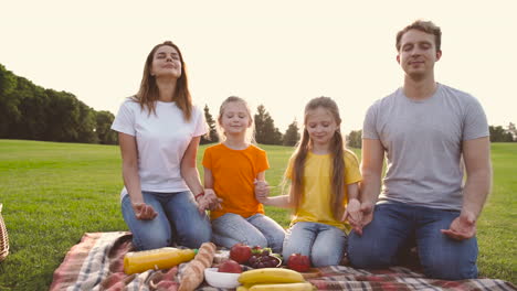 Happy-Parents-And-Daughters-With-Closed-Eyes-Kneeling-On-Meadow,-Holding-Hands-And-Meditating-During-A-Picnic-In-The-Park-1