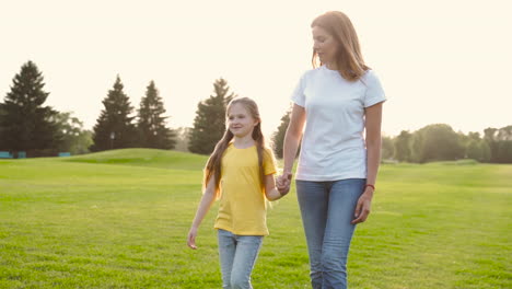 Mother-And-Little-Daughter-Holding-Hands-And-Talking-While-Walking-On-Green-Grass-Field-In-The-Park