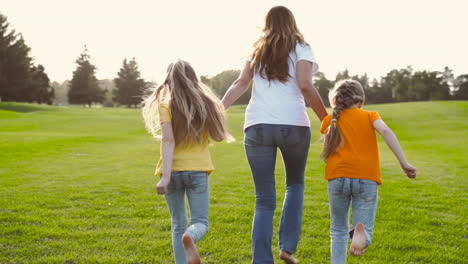 Back-View-Of-Mother-And-Her-Two-Little-Daughters-Holding-Hands-And-Running-On-Green-Grass-Field-In-The-Park