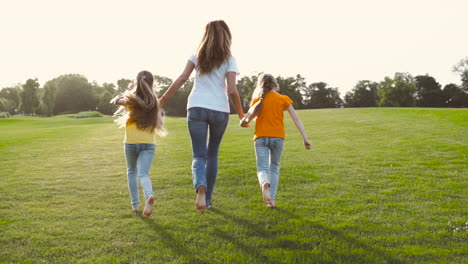 Back-View-Of-Mother-And-Her-Two-Little-Daughters-Holding-Hands-And-Running-On-Green-Grass-Field-In-The-Park-1