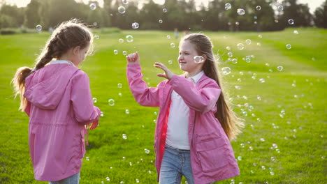 Happy-Little-Sisters-In-Identical-Clothes-Catching-Soap-Bubbles,-Holding-Hands-And-Spinning-Around-In-The-Park