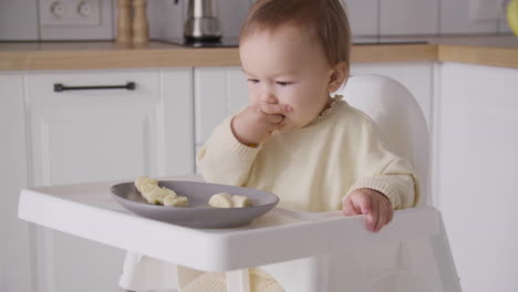 Cute-Baby-Girl-Eating-Banana-Slices-Sitting-In-Her-High-Chair-In-The-Kitchen-1