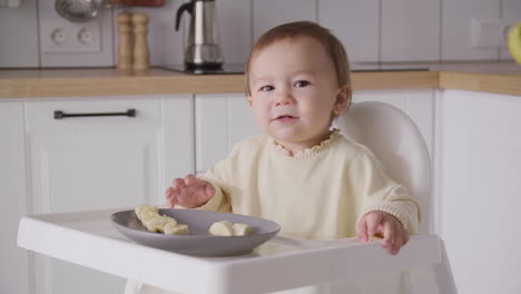 Cute-Baby-Girl-Eating-Banana-Slices-Sitting-In-Her-High-Chair-In-The-Kitchen-2