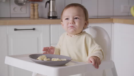Cute-Baby-Girl-Eating-Banana-Slices-Sitting-In-Her-High-Chair-In-The-Kitchen-3