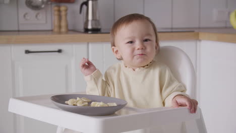 Cute-Baby-Girl-Eating-Banana-Slices-Sitting-In-Her-High-Chair-In-The-Kitchen-4