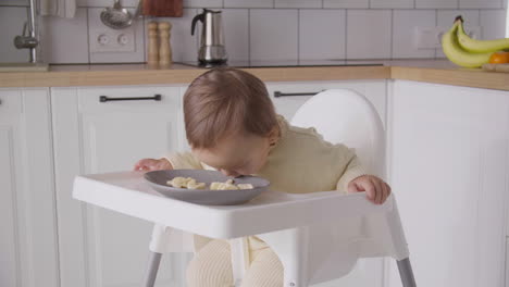 Cute-Baby-Girl-Eating-Banana-Slices-Sitting-In-Her-High-Chair-In-The-Kitchen-5