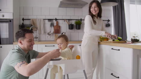 Happy-Father-Feeding-His-Cute-Baby-Girl-Sitting-In-Her-High-Chair-In-The-Kitchen-While-Smiling-Mother-Cutting-Fruit-And-Looking-At-Them