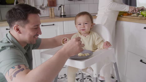 Happy-Father-Feeding-His-Cute-Baby-Girl-Sitting-In-Her-High-Chair-In-The-Kitchen-While-Mother-Cutting-Fruit-Behind-Them