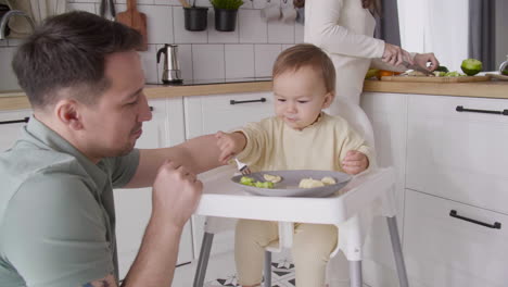 Father-Feeding-His-Happy-Baby-Girl-Sitting-In-Her-High-Chair-In-The-Kitchen-While-Mother-Cutting-Fruit-Behind-Them