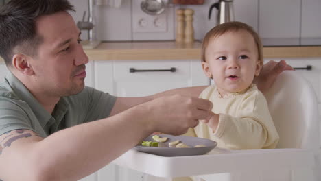 Happy-Father-Feeding-His-Cute-Baby-Girl-Sitting-In-Her-High-Chair-In-The-Kitchen