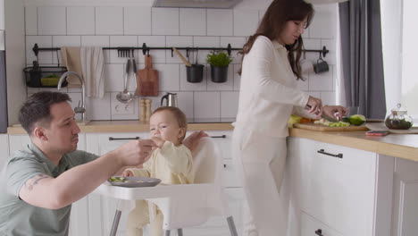 Father-Feeding-His-Happy-Baby-Girl-Sitting-In-Her-High-Chair-In-The-Kitchen-While-His-Wife-Cutting-Fruit-And-Talking-With-Him