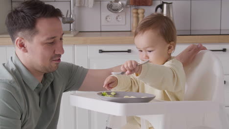 Cute-Baby-Girl-Sitting-In-High-Chair,-Holding-Fork-And-Playing-With-Food-While-Her-Father-Looking-At-Her