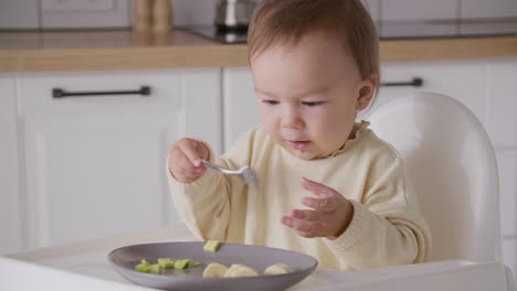 Cute-Baby-Girl-Eating-Avocado-Slices-Sitting-In-Her-High-Chair-In-The-Kitchen