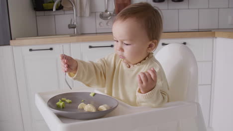 Cute-Baby-Girl-Eating-Avocado-Slices-Using-Fork-While-Sitting-In-Her-High-Chair-In-The-Kitchen