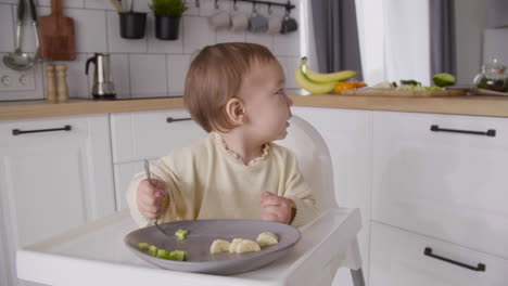 Cute-Baby-Girl-Holding-Fork-And-Looking-Around-While-Sitting-In-Her-High-Chair-In-The-Kitchen-And-Eating-Fruit