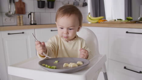 Cute-Baby-Girl-Holding-Fork-And-Trying-To-Eat-Avocado-Slices-While-Sitting-In-Her-High-Chair-In-The-Kitchen