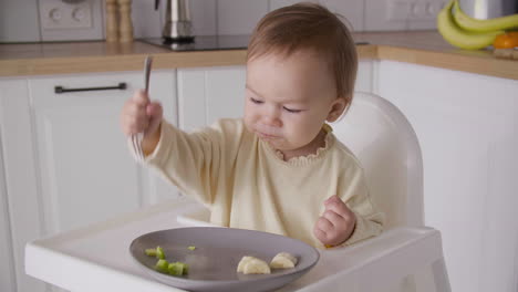Cute-Baby-Girl-Sitting-In-High-Chair-In-The-Kitchen-And-Holding-Fork-And-Eating-Fruit-Slices