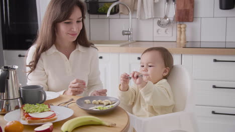Happy-Mother-Feeding-Her-Cute-Little-Girl-Sitting-In-High-Chair-In-The-Kitchen