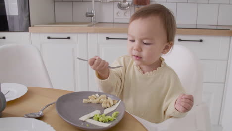 Cute-Baby-Girl-Sitting-In-High-Chair-Next-To-The-Kitchen-Table-Eating-Food-With-Fork-And-Hands