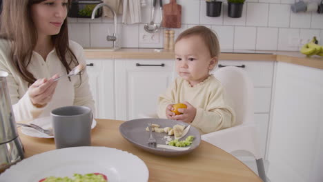Cute-Little-Girl-Sitting-In-High-Chair-In-The-Kitchen-And-Holding-A-Clementine-While-Her-Mom-Feeding-Her