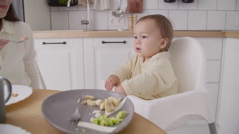 Cute-Little-Girl-Sitting-In-High-Chair-In-The-Kitchen-While-Her-Mother-Feeding-Her-2