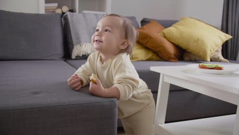 Cute-Little-Girl-Standing-Next-To-Sofa-At-Living-Room-And-Eating-Bread