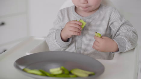 Close-Up-Of-A-Cute-Baby-Boy-Eating-Avocado-Slices-Sitting-In-High-Chair-In-The-Kitchen