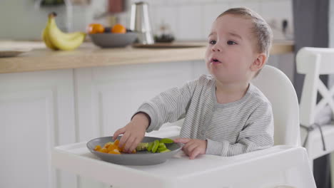 Cute-Baby-Boy-Eating-Clementine-Sitting-In-High-Chair-In-The-Kitchen