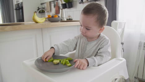 Cute-Baby-Boy-Eating-Clementine-And-Avocado-Sitting-In-High-Chair-In-The-Kitchen