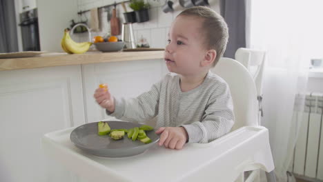 Cute-Baby-Boy-Eating-Clementine-And-Avocado-Sitting-In-High-Chair-In-The-Kitchen-1