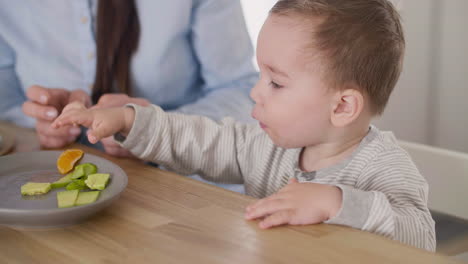 Cute-Baby-Boy-Taking-Segment-Of-Clementine-From-Plate-On-Table-While-His-Mom-Sitting-Next-To-Him