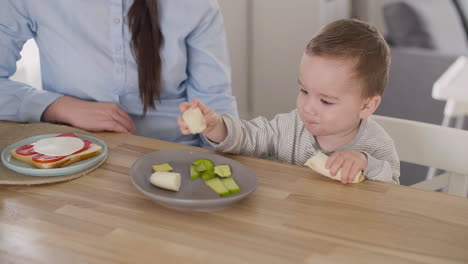 Cute-Baby-Boy-Taking-Banana-From-Plate-And-Eating-While-His-Mom-Sitting-Next-To-Him-At-Home