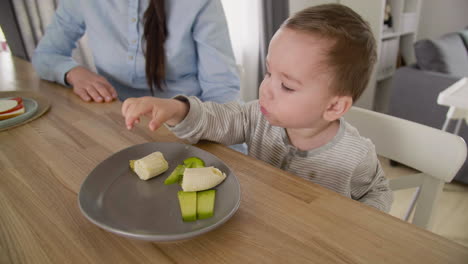 Cute-Baby-Boy-Playing-With-Food-While-Sitting-At-Table-With-His-Mother-In-Living-Room