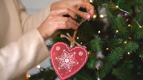 Vista-De-Cerca-De-La-Mano-De-La-Mujer-Colgando-Un-Corazón-Rojo-De-Madera-En-El-árbol-De-Navidad