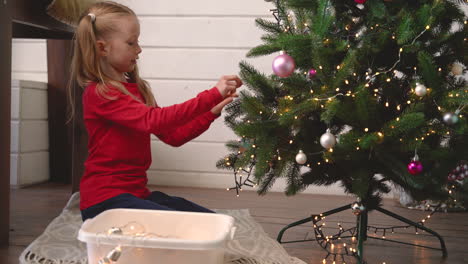 Kleines-Blondes-Mädchen,-Das-Auf-Dem-Boden-Kniet-Und-Weihnachtsdekoration-Aus-Einer-Kiste-Aufhebt-Und-An-Den-Weihnachtsbaum-Hängt
