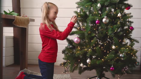 Little-Blonde-Girl-De-Kneeling-On-The-Floor-Hanging-Christmas-Decoration-On-Christmas-Tree