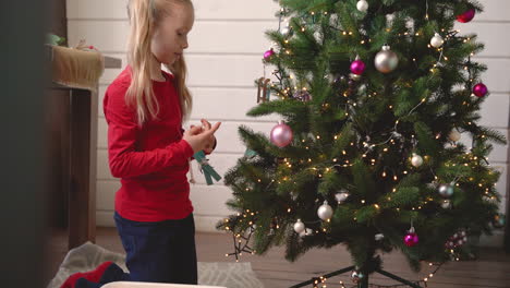 Kleines-Blondes-Mädchen,-Das-Auf-Dem-Boden-Kniet,-Weihnachtsdekoration-Aus-Einer-Kiste-Aufhebt-Und-An-Den-Weihnachtsbaum-Hängt-1