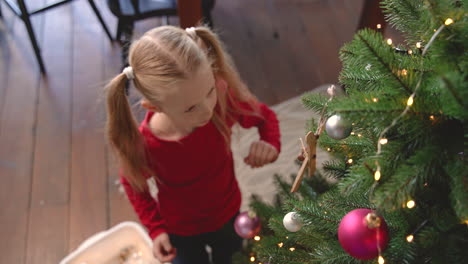 Blick-Von-Oben-Auf-Das-Kleine-Blonde-Mädchen,-Das-Auf-Dem-Boden-Kniet-Und-Weihnachtsdekoration-Aus-Einer-Kiste-Aufhebt-Und-An-Den-Weihnachtsbaum-Hängt