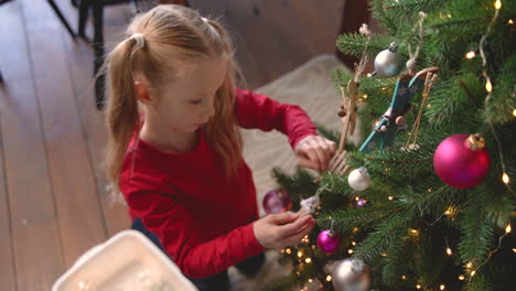 Blick-Von-Oben-Auf-Das-Kleine-Blonde-Mädchen,-Das-Auf-Dem-Boden-Kniet-Und-Weihnachtsdekoration-Aus-Einer-Kiste-Aufhebt-Und-An-Den-Weihnachtsbaum-Hängt-1