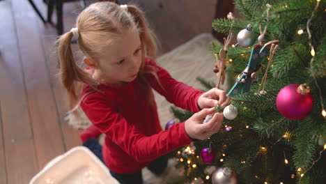 Top-View-Of-Little-Blonde-Girl-De-Kneeling-On-The-Floor-Picking-Up-Christmas-Decoration-From-A-Box-And-Hanging-It-On-Christmas-Tree-2