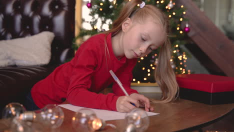 Blondes-Mädchen,-Das-Am-Tisch-Sitzt-Und-Auf-Dem-Papier-Neben-Dem-Weihnachtsbaum-Schreibt