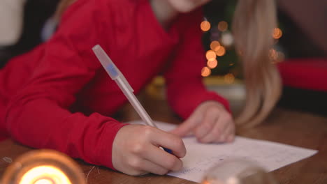 Close-Up-View-Of-Blonde-Girl-Sitting-At-The-Table-Writing-On-The-Paper-Near-The-Christmas-Tree