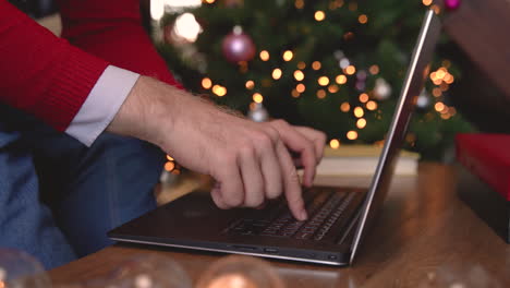 Close-Up-View-Of-Man-Hands-Typing-On-Laptop-Sitting-Near-The-Christmas-Tree-In-Living-Room-With-Christmas-Decoration
