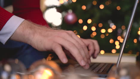 Nahaufnahme-Von-Mannhänden,-Die-Auf-Einem-Laptop-Tippen,-Der-Neben-Dem-Weihnachtsbaum-Im-Wohnzimmer-Mit-Weihnachtsdekoration-Sitzt-2